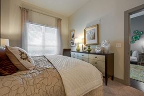 Dallas apartment for rent - A bedroom here at Cityplace Heights with a neatly made bed in neutral bedding, a dark dresser with green and white decor, framed wall art, a table lamp, and a window with blinds and sheer curtains.