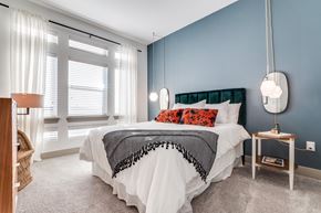 Farmers Branch apartment for rent - A bright bedroom here at Crosby at the Brickyard with a blue accent wall, plush white bed, bold orange pillows, and large windows, creating a cozy, modern, inviting space.