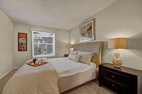 Midland apartment for rent - A comfortable bedroom here at Cypress Pointe with plush beige carpet, neutral walls, and a large window with vertical blinds allowing natural light to fill the room. A neatly staged bed with soft bedding and matching bedside lamps creates a calm, cozy, and inviting atmosphere.