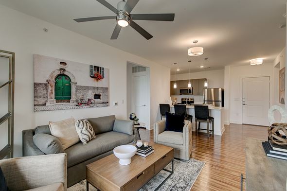 Rockwall apartment for rent - A bright open living room and kitchen here at Florence at the Harbor with wood-style flooring, a gray sofa, accent chairs, ceiling fan, breakfast bar seating, and stainless steel appliances, creating a modern, welcoming feel.