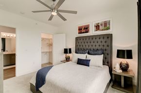 Rockwall apartment for rent - A comfortable bedroom here at Florence at the Harbor with a tufted headboard, plush carpeting, ceiling fan, bedside lamps, and a walk-in closet with attached bathroom, creating a calm and inviting retreat.