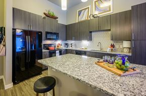 Rowlett apartment for rent - A modern kitchen here at Harmony Luxury with dark wood cabinetry, granite countertops, and a large center island, featuring sleek black appliances, pendant lighting, a patterned tile backsplash, and bar-style seating that create a polished, upscale atmosphere.