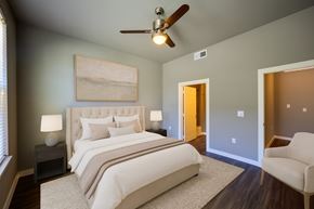 Rowlett apartment for rent - A serene bedroom here at Harmony Luxury with a plush upholstered bed, soft neutral bedding, and matching bedside tables, complemented by a ceiling fan, warm accent lighting, and an adjoining bathroom entry that creates a calm, upscale retreat.
