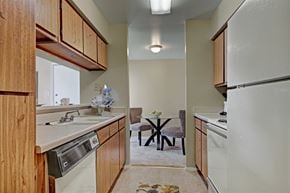 Odessa apartment for rent - A narrow kitchen here at High Plains with warm wood cabinets, light countertops, and white appliances, featuring a dishwasher and a compact layout that opens to a small dining area, creating a simple, functional, and comfortable cooking space.