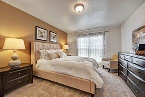 Odessa apartment for rent - A welcoming bedroom here at High Plains with plush beige carpet, a neutral upholstered bed, warm accent walls, and soft lighting, complemented by a large window with patterned curtains and tasteful furnishings that create a calm, comfortable retreat.