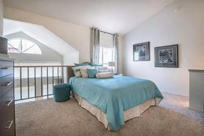 Dallas apartment for rent - A bedroom here at Lakewood on the Trail with soft carpet, a teal bedspread and matching accent pillows, a small teal ottoman, and bright windows near a loft-style railing, creating a fresh and inviting sleeping space.