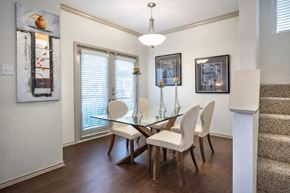 Dallas apartment for rent - A dining area here at Lakewood on the Trail with a glass-top table, four cream upholstered chairs, silver candle holders, framed wall art, dark wood floors, and bright patio doors with white blinds.