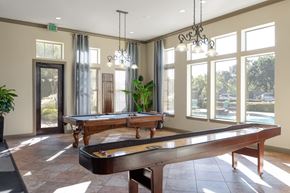Mansfield apartment for rent - A game room here at Landing at Mansfield with a wooden pool table and shuffleboard table, tall bright windows, dark metal chandeliers, and warm tile flooring overlooking the pool area.