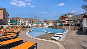 Odessa apartment for rent - A resort-style outdoor pool here at Latitude 31 featuring a curved swimming pool with in-water lounge chairs, surrounding sun deck lined with orange chaise loungers, shaded seating areas with umbrellas, cascading water features, and upscale apartment buildings creating a vibrant and relaxing community atmosphere.