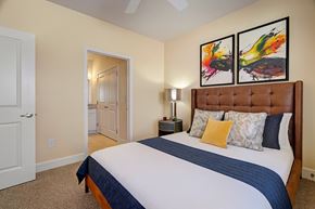 Odessa apartment for rent - A stylish primary bedroom here at Latitude 31 Apartments featuring a plush upholstered bed with crisp bedding and layered accent pillows, soft neutral walls, cozy carpet flooring, modern abstract wall art above the headboard, and a private doorway leading to an en suite bathroom that enhances comfort and convenience.