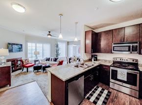 Midland apartment for rent - A bright open-concept living room here at Le Mirage featuring an adjoining kitchen with dark wood cabinetry, stone-look countertops, and pendant lighting, a cozy seating area with neutral sofa and accent chairs, large windows with vertical blinds for natural light, soft carpeting, and modern finishes throughout.