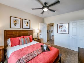 Midland apartment for rent - A cozy primary bedroom here at Le Mirage featuring a wooden bed frame with coral bedding and layered pillows, soft carpet flooring, a ceiling fan with light, matching bedside tables with lamps, framed wall art, and an open walk-in closet with shelving and hanging space visible through a white door.