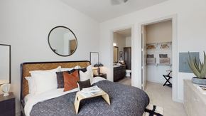 Allen apartment for rent - A bright bedroom here at Magnolia on the Green with plush carpet, a large bed with layered pillows, bedside lamps, a round wall mirror, and an open closet and bathroom doorway for a comfortable, connected layout.