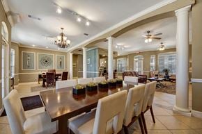 Odessa apartment for rent - A spacious clubhouse interior here at Mission Green featuring a large dining table with upholstered chairs, decorative columns and archways, elegant chandeliers and ceiling fans, tiled flooring, and adjacent lounge seating with sofas and coffee tables, creating a welcoming shared space for dining, relaxing, and gathering with neighbors.
