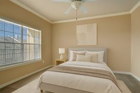 Odessa apartment for rent - A cozy and serene bedroom here at Mission Green featuring a neatly made bed with white bedding, layered neutral pillows, and a soft throw blanket, complemented by warm beige walls, plush carpeting, a ceiling fan with light, simple abstract wall art above the headboard, and natural light filtering through wide window blinds.