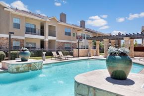 Odessa apartment for rent - A resort-style outdoor pool area here at Mission Green showcasing clear blue water, a spacious stone pool deck, large decorative planters, shaded pergola columns, lounge chairs, and surrounding apartment buildings under a bright sky, creating a relaxing community space for residents to unwind.