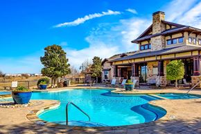 Fort Worth apartment for rent - An outdoor pool area here at Monterra Village with a curved blue pool, tan stone deck, large blue planters, lounge chairs, and a stone clubhouse with covered patio seating under a bright sky.