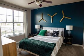 Fort Worth apartment for rent - A cozy bedroom here at Monterra Village with soft carpet, a large bed with green and neutral bedding, a bold blue accent wall with decorative art, bedside lamps, and a bright window with blinds.