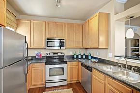 Midland apartment for rent - A bright, modern kitchen here at Palms at Briarwood featuring light wood cabinetry, dark stone countertops, a stainless steel refrigerator, oven, microwave, and dishwasher, a double-basin sink, and warm wood-style flooring, with ample cabinet storage and an open layout that connects seamlessly to the adjacent living space.