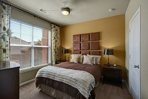 Midland apartment for rent - A cozy bedroom here at Palms at Briarwood featuring a comfortable bed with layered bedding, matching nightstands with lamps, soft carpet flooring, and a large window with blinds and patterned curtains that bring in natural light while creating a warm, inviting retreat.