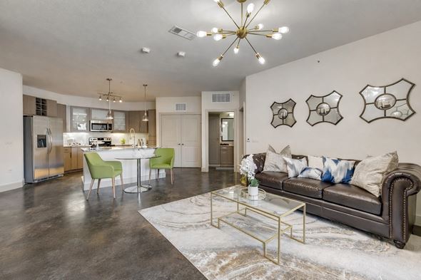Dallas apartment for rent - A modern open living and kitchen area here at Skyline Farmers Market with a brown leather sofa, green dining chairs, glass coffee table, light wood cabinets, and a bright chandelier over polished concrete floors.