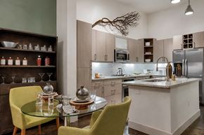 Dallas apartment for rent - A dining and kitchen area here at Skyline Farmers Market with light wood cabinets, stainless appliances, a white island, glass round table, and standout green dining chairs beside a dark wood display shelf.