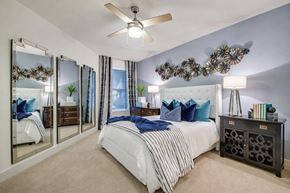 Dallas apartment for rent - A bedroom here at Skyline Farmers Market with a white tufted bed, blue and teal accent pillows, navy throw blanket, mirrored wall panels, black nightstands, and soft neutral carpet under bright lighting.