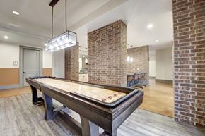 Fairview apartment for rent - A game lounge here at Sorrel Fairview with a shuffleboard table, brick columns, warm concrete floors, modern rectangular pendant light, and a bar-height counter with stools in the background.