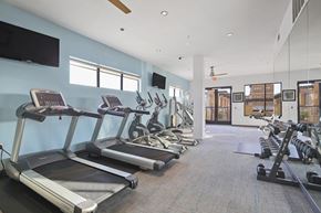 Dallas apartment for rent - A fitness center here at The Uptown with modern cardio machines, treadmills and ellipticals, a mirrored wall with dumbbell rack, blue accent wall, and bright natural light from tall windows.