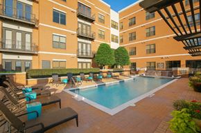 Dallas apartment for rent - A pool courtyard here at The Uptown with a rectangular swimming pool, brick deck, black lounge chairs, bright teal side tables, trimmed green trees, and surrounding mid-rise brick apartment buildings in sunny light.