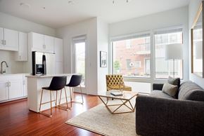 Dallas apartment for rent - A bright living room here at The Uptown with a charcoal sofa, gold tufted accent chair, black bar stools at a white kitchen island, large courtyard-facing windows, and warm wood floors in an open layout.