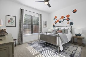 Dallas apartment for rent - A bedroom here at The Uptown with a gray metal bed frame, white bedding, colorful orange and teal accent pillows, geometric wall art, patterned gray rug, wood nightstand, and bright window with striped curtains.