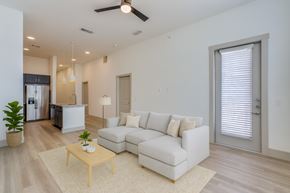 Lewisville apartment for rent - A living room here at Valor at the Realm with a light gray sectional sofa, light wood coffee table, cream rug, small green plants, and an open kitchen with dark cabinets and stainless appliances in back.