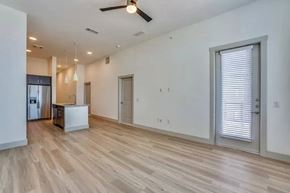 Lewisville apartment for rent - A bright open-concept living and kitchen area here at Valor at the Realm with light wood flooring, white walls, a modern island with pendant lights, stainless steel refrigerator, and a glass door with blinds.
