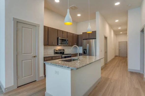 Lewisville apartment for rent - A kitchen here at Valor at the Realm with dark brown cabinets, stainless steel appliances, a granite island with sink, white pendant lights, and light wood flooring leading into a long hallway.