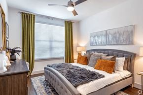 Lewisville apartment for rent - A bedroom here at Valor at the Realm with a gray tufted bed, layered white and gray bedding, rust accent pillows, green curtains, dark wood dresser, patterned rug, and warm bedside lamps for a cozy, polished look.