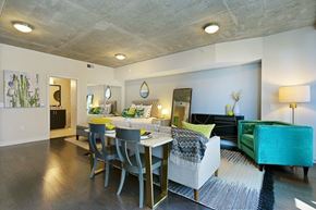 Dallas apartment for rent - A modern open studio here at Victory Place with dark wood flooring, a dining table, cozy seating, green accent furniture, and a sleeping area with decorative bedding and nearby bathroom access.