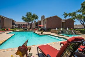 Midland apartment for rent - A resort-style outdoor pool here at Wildflower featuring clear blue water, red cushioned lounge chairs, shaded umbrellas, and a stone water feature, surrounded by landscaped greenery and apartment buildings, creating a relaxing and inviting community space for residents to enjoy warm days outdoors.