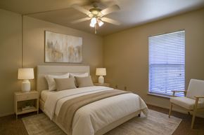 Midland apartment for rent - A calm, modern bedroom here at Wildflower featuring a neutral upholstered bed with crisp white bedding, textured accent pillows, and a soft throw blanket. Coordinated nightstands with matching lamps, a ceiling fan with lighting, plush carpeting, and a large window with blinds create a comfortable, restful retreat filled with natural light.