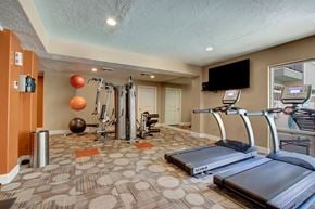 Salt Lake City apartment for rent - A fitness room here at Bridges with treadmills, a cable weight machine, wall mirrors, mounted TV, patterned carpet, and orange accent walls with exercise balls and strength equipment.