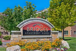 Salt Lake City apartment for rent - A community monument sign here at Bridges with a stone base and curved red-and-black Bridges Apartment Homes panel, surrounded by bright yellow flowers, green trees, and landscaped greenery under a clear blue sky.