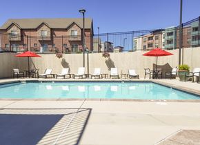 Sandy apartment for rent - A pool area here at Hills at Sandy Station with a clear blue swimming pool, white lounge chairs, and red patio umbrellas along a fenced concrete deck under bright sunlight.