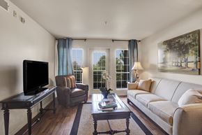 Sandy apartment for rent - A living room here at Legends at River Oaks with a light neutral sofa, brown leather accent chair, blue curtains, glass coffee table, dark TV console, and bright balcony doors bringing in natural light.