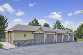 Sandy apartment for rent - A detached garage building here at Legends at River Oaks with multiple gray garage doors, tan and stone exterior walls, dark asphalt drive, and green trees behind under a bright blue sky.