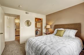Sandy apartment for rent - A bedroom here at Legends at River Oaks with a white quilted bedspread, woven headboard, tan accent wall, open closet, round wall mirror, soft carpet, and a warm bedside lamp.