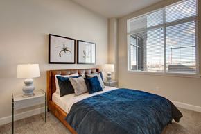 Salt Lake City apartment for rent - A bright bedroom here at Ritz Classic with a warm brown upholstered bed frame, blue bedding and accent pillows, matching nightstands with lamps, soft carpet, and a large window with blinds bringing in natural light and city views.