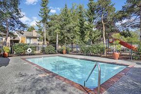Lynnwood apartment for rent - A pool area here at A'Cappella with a clear blue swimming pool, brick trim deck, terracotta planters, black fencing, and nearby playground, creating a bright, relaxed, and family-friendly outdoor space.