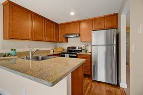 Bellevue apartment for rent - A modern kitchen here at Adagio featuring warm wood cabinetry, granite-style countertops, and a peninsula with a built-in sink and curved faucet, complemented by stainless steel appliances, a range hood, and wood-look flooring, while recessed lighting and an open layout provide a clean, functional space with ample storage and prep areas.