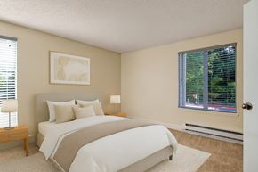 Bellevue apartment for rent - A calm, inviting bedroom here at Adagio featuring plush beige carpeting, soft neutral walls, and a comfortable bed with layered white and neutral bedding, complemented by matching wood bedside tables and lamps, while large windows with blinds allow natural light and offer peaceful greenery views outside.