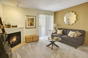 Covington apartment for rent - A stylish living room here at Allegro featuring plush neutral carpeting, a comfortable gray sofa with patterned accent pillows, and a round glass coffee table centered in the space, while a corner fireplace adds warmth, decorative wall art enhances the room, and a sliding glass door with vertical blinds allows natural light to fill the inviting layout.